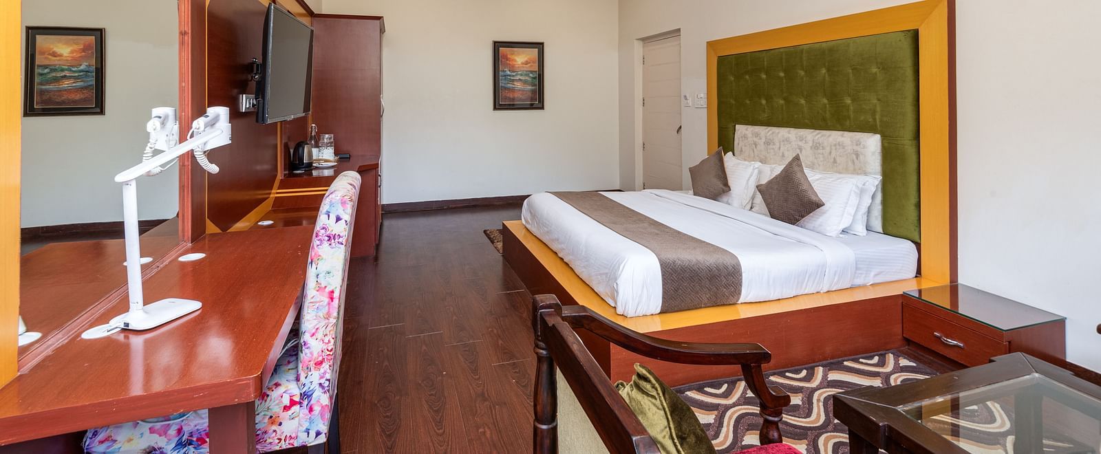 The image shows a spacious hotel room with a large bed, seating area, and desk. It's decorated with artwork and has a ceiling fan | DLS Tehri Club Resort